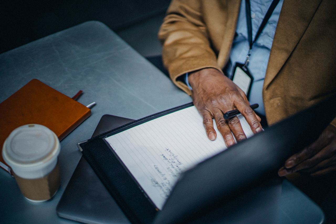 From above of crop anonymous ethnic male executive with paper sheet in folder on table with takeaway coffee in cafeteria